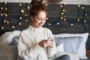 Jeune fille sur son téléphone portable assise sur son lit avec une guirlande lumineuse en fond